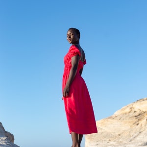 May include: A woman wearing a bright red dress stands on a rocky surface with a blue sky in the background.