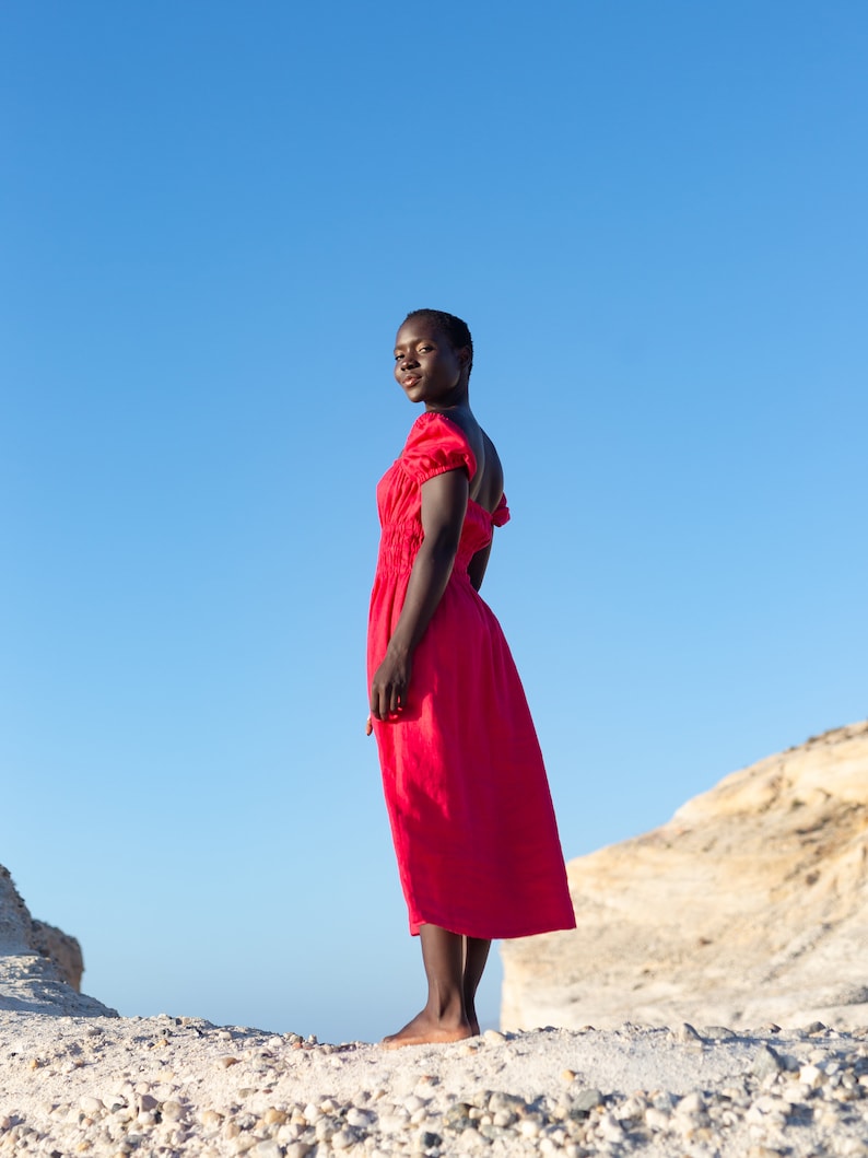 May include: A woman wearing a bright red dress stands on a rocky surface with a blue sky in the background.