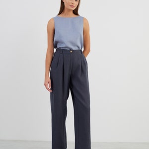 May include: A woman wearing a light blue sleeveless top and dark blue high-waisted trousers with pleats. She is wearing black loafers with gold buckles.