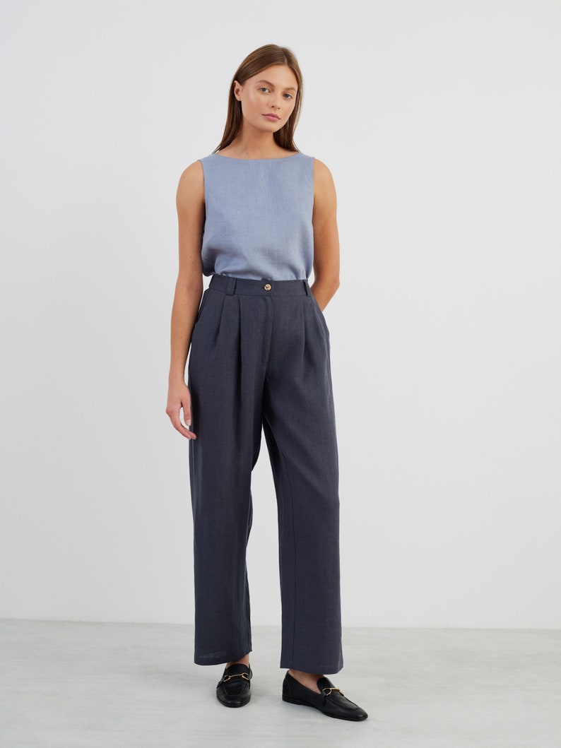 May include: A woman wearing a light blue sleeveless top and dark blue high-waisted trousers with pleats. She is wearing black loafers with gold buckles.