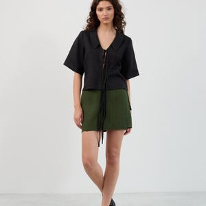 K&ouml;nnte beinhalten: Ein schwarzes Oberteil mit kurzen &Auml;rmeln, Kragen und Bindeband, kombiniert mit einem gr&uuml;nen Minirock. Das Outfit wird durch schwarze Loafer vervollst&auml;ndigt. Die Kleidung ist aus einem strukturierten Stoff gefertigt.