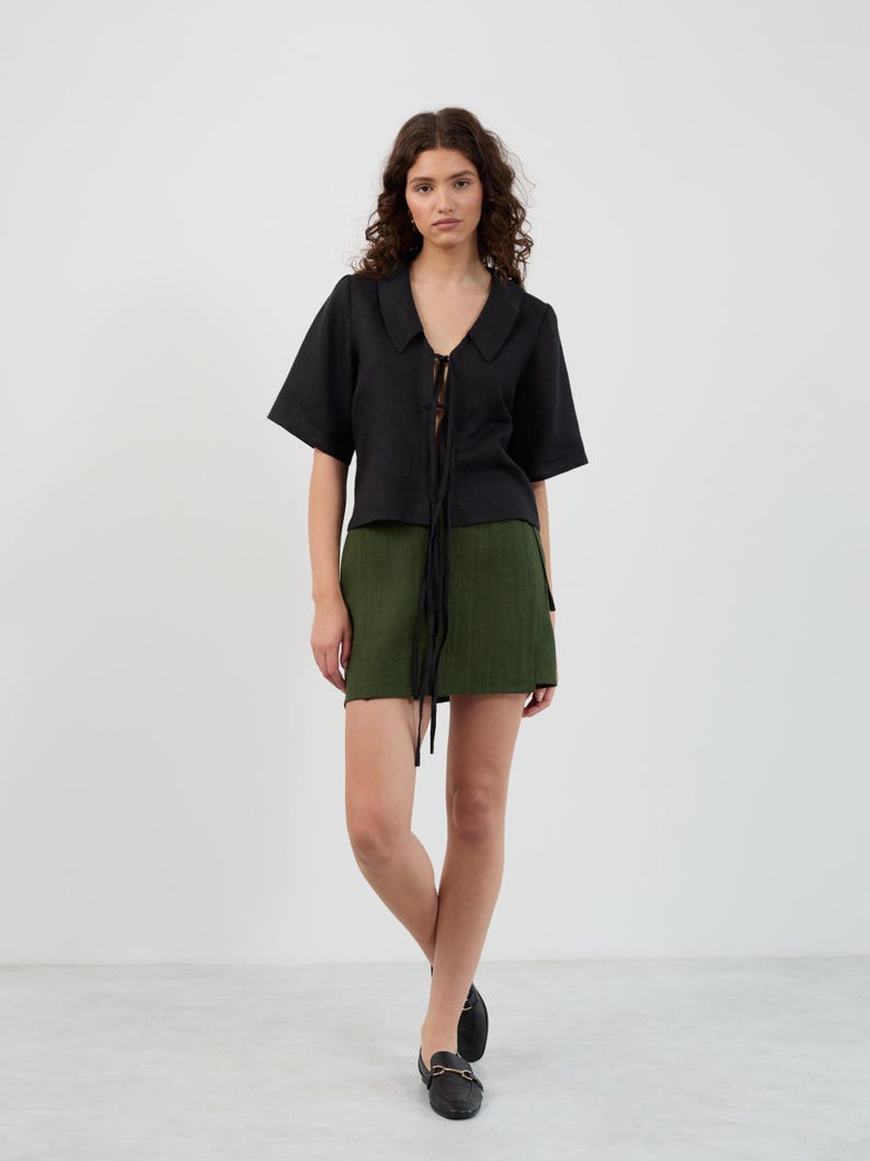 K&ouml;nnte beinhalten: Ein schwarzes Oberteil mit kurzen &Auml;rmeln, Kragen und Bindeband, kombiniert mit einem gr&uuml;nen Minirock. Das Outfit wird durch schwarze Loafer vervollst&auml;ndigt. Die Kleidung ist aus einem strukturierten Stoff gefertigt.