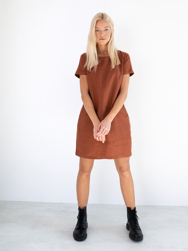 May include: A woman wearing a rust-colored linen dress with short sleeves and a square neckline. She is standing on a white background and wearing black lace-up boots.