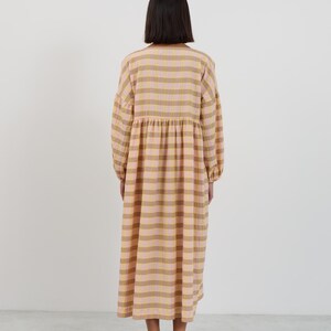 K&ouml;nnte beinhalten: Ein lang&auml;rmeliges Midikleid mit einem karierten Muster in Hellrosa und Hellbraun. Das Kleidungsst&uuml;ck hat eine geraffte Taille, einen Rundhalsausschnitt und einen lockeren, flie&szlig;enden Rock, der unter die Knie reicht. Der Stoff hat eine leicht knittrige Textur.
