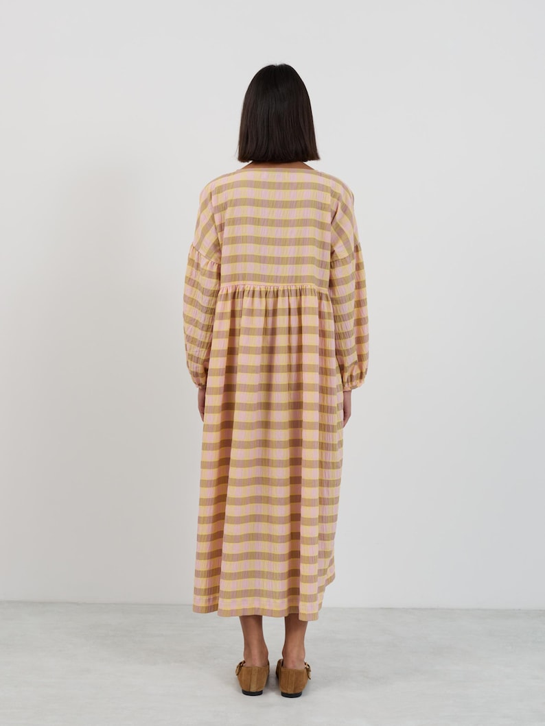 K&ouml;nnte beinhalten: Ein lang&auml;rmeliges Midikleid mit einem karierten Muster in Hellrosa und Hellbraun. Das Kleidungsst&uuml;ck hat eine geraffte Taille, einen Rundhalsausschnitt und einen lockeren, flie&szlig;enden Rock, der unter die Knie reicht. Der Stoff hat eine leicht knittrige Textur.
