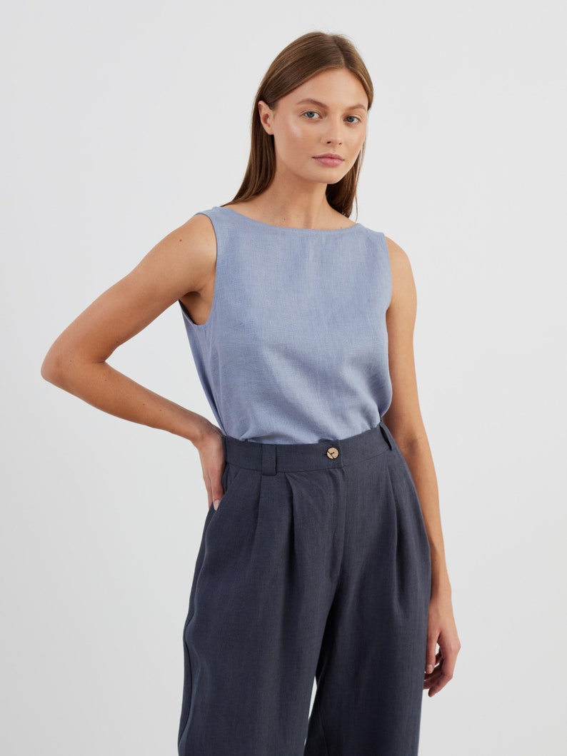 May include: A woman wearing a light blue sleeveless top and dark grey wide-leg trousers. The trousers have a button fastening and pleats at the front.
