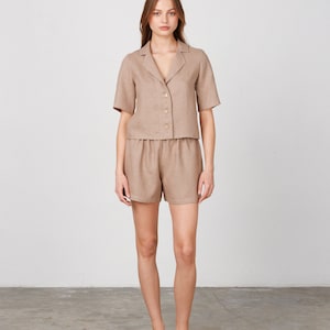 May include: A woman is wearing a light brown linen short-sleeved shirt and matching shorts. The shirt has a collar and buttons down the front. The shorts have a drawstring waist.