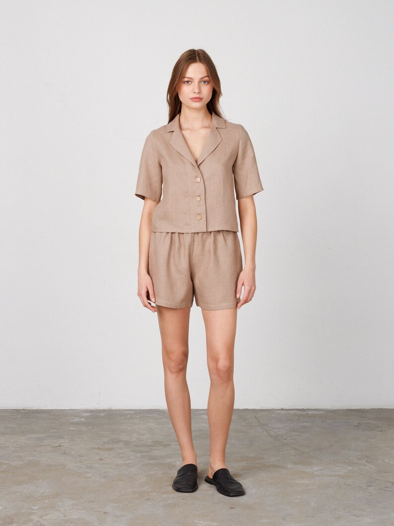 May include: A woman is wearing a light brown linen short-sleeved shirt and matching shorts. The shirt has a collar and buttons down the front. The shorts have a drawstring waist.