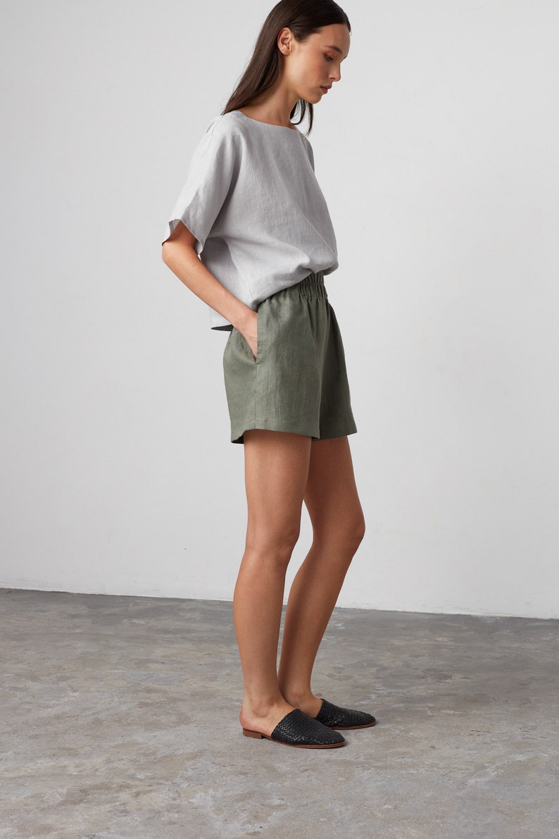 Pode incluir: Uma mulher vestindo uma blusa de linho cinza claro e shorts de linho verde oliva. Ela est&aacute; em p&eacute; em um piso de concreto cinza e usando sand&aacute;lias de couro tran&ccedil;ado preto.