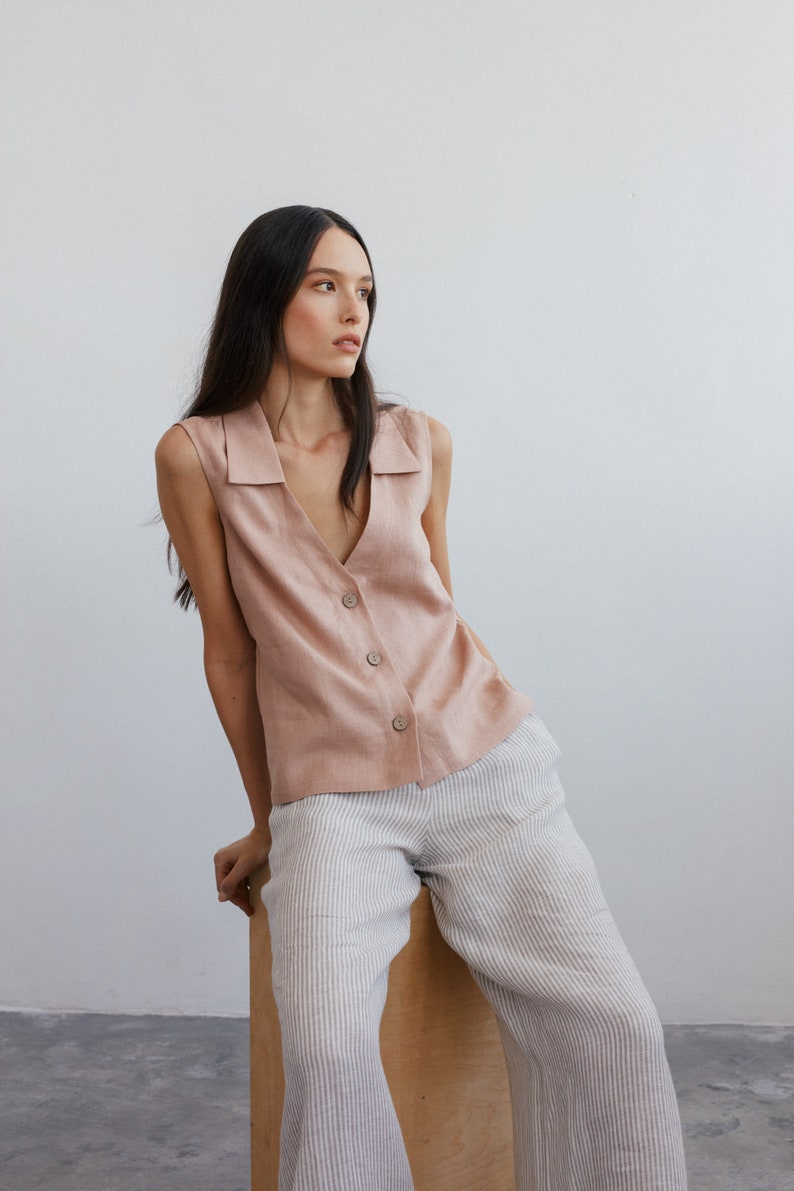 Pu&ograve; includere: Una donna indossa una camicetta di lino rosa chiaro senza maniche con colletto e chiusura con bottoni. Indossa anche pantaloni a righe.