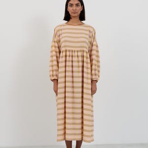 K&ouml;nnte beinhalten: Ein midilanges Kleid mit einem Karomuster in Hellrosa und ged&auml;mpftem Olivgr&uuml;n. Das Kleidungsst&uuml;ck hat einen Rundhalsausschnitt, lange &Auml;rmel mit gerafften B&uuml;ndchen und eine geraffte Taille, die eine lockere, flie&szlig;ende Silhouette bis zum Saum erzeugt.