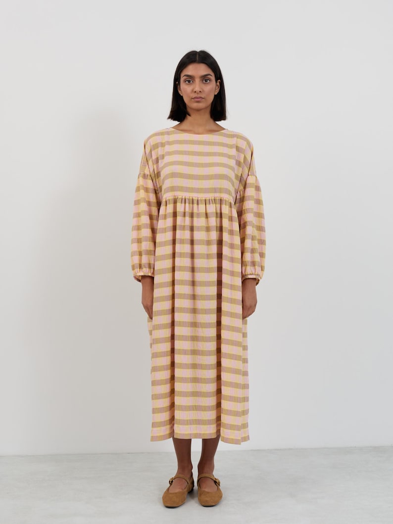 K&ouml;nnte beinhalten: Ein midilanges Kleid mit einem Karomuster in Hellrosa und ged&auml;mpftem Olivgr&uuml;n. Das Kleidungsst&uuml;ck hat einen Rundhalsausschnitt, lange &Auml;rmel mit gerafften B&uuml;ndchen und eine geraffte Taille, die eine lockere, flie&szlig;ende Silhouette bis zum Saum erzeugt.