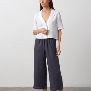 May include: A white linen shirt with a collar and short sleeves. The shirt is buttoned up and has a relaxed fit. The model is wearing dark gray linen pants and black leather flats.