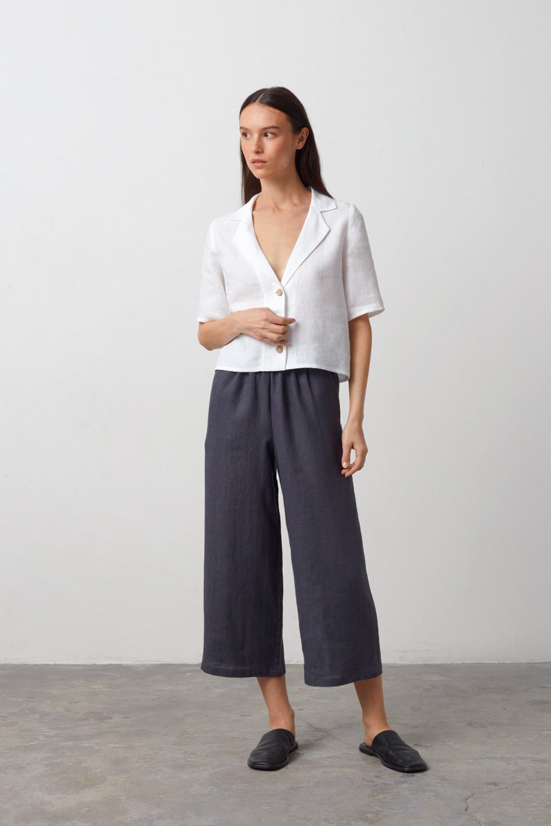 May include: A white linen shirt with a collar and short sleeves. The shirt is buttoned up and has a relaxed fit. The model is wearing dark gray linen pants and black leather flats.