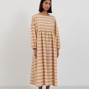 K&ouml;nnte beinhalten: Ein lang&auml;rmeliges Midikleid mit einem horizontalen Karomuster in sanften T&ouml;nen von blassem Rosa und ged&auml;mpftem Senfgelb. Das Kleidungsst&uuml;ck hat einen Rundhalsausschnitt, eine geraffte Taille und locker sitzende &Auml;rmel mit elastischen B&uuml;ndchen.