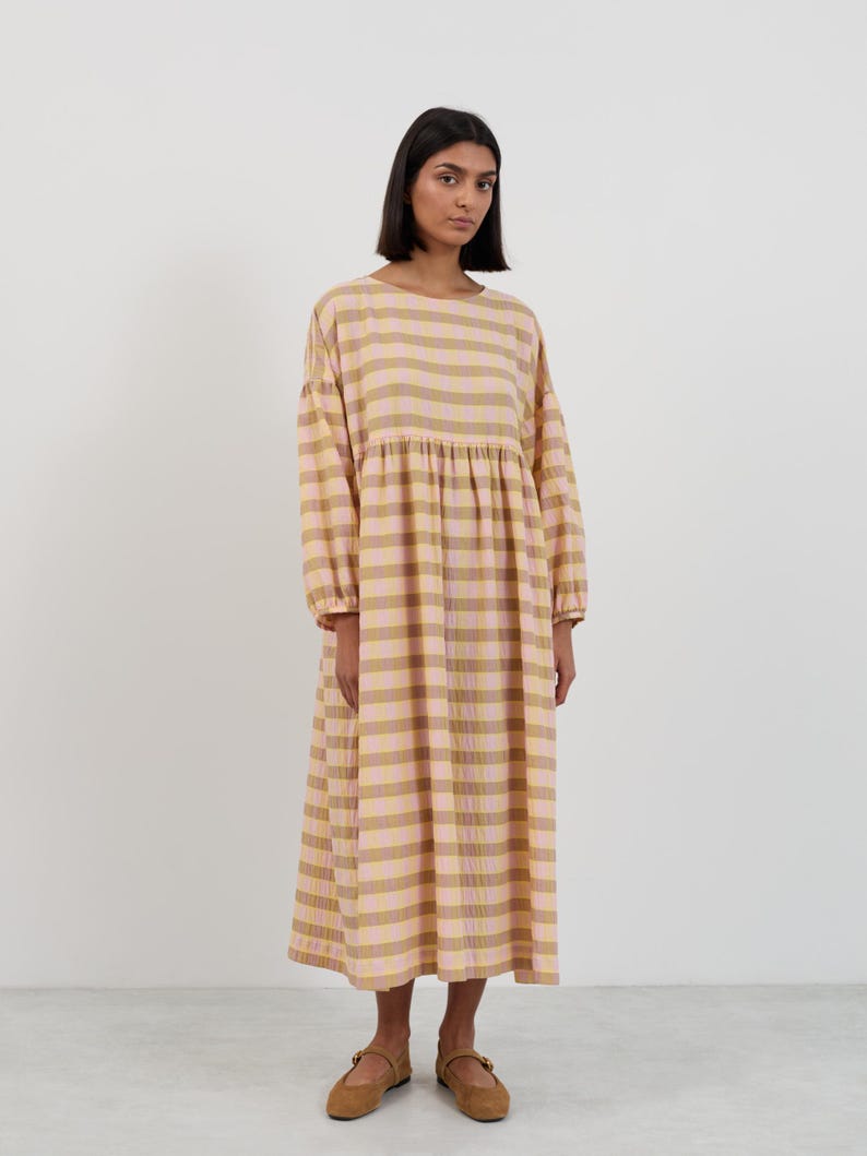 K&ouml;nnte beinhalten: Ein lang&auml;rmeliges Midikleid mit einem horizontalen Karomuster in sanften T&ouml;nen von blassem Rosa und ged&auml;mpftem Senfgelb. Das Kleidungsst&uuml;ck hat einen Rundhalsausschnitt, eine geraffte Taille und locker sitzende &Auml;rmel mit elastischen B&uuml;ndchen.