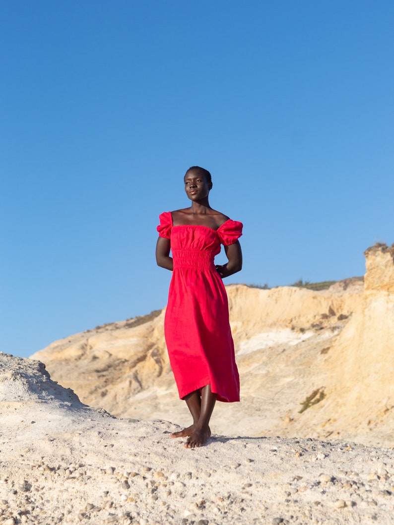 May include: A person wearing a red dress stands on a sandy hill with a blue sky in the background.