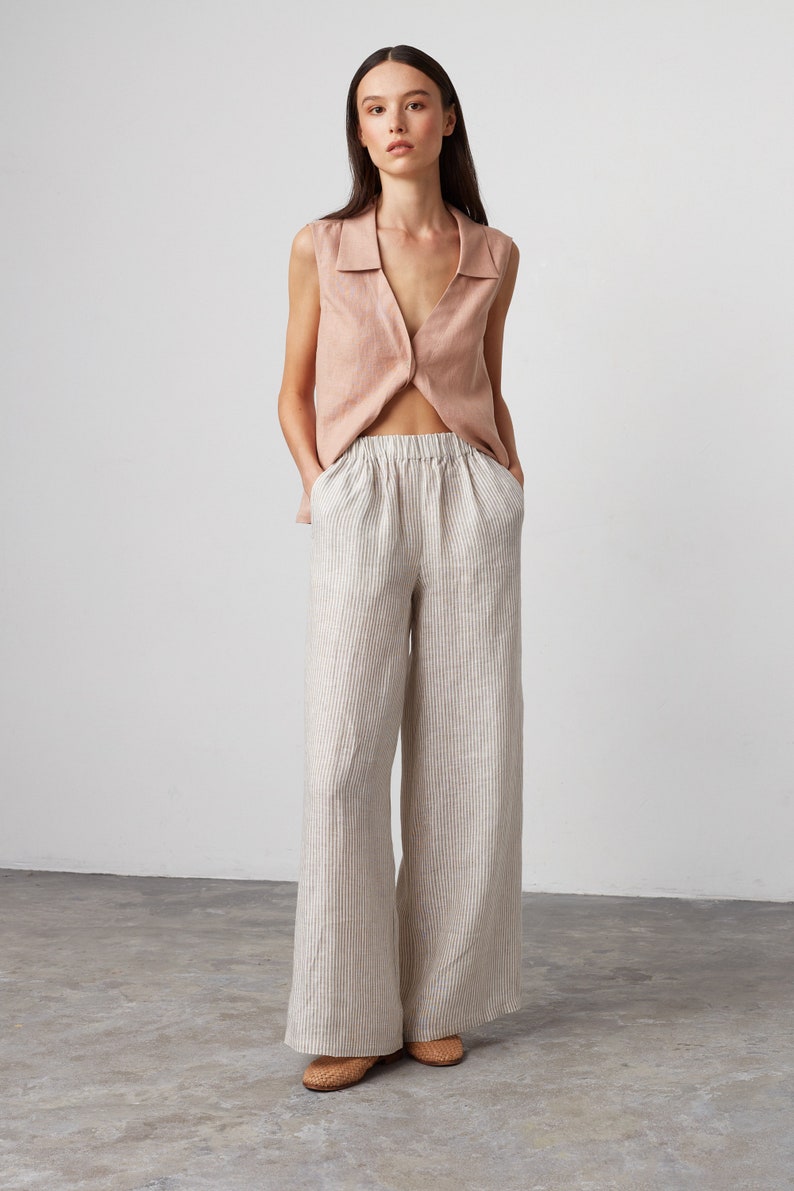 Pu&ograve; includere: Una donna indossa una camicetta rosa chiaro senza maniche con colletto e pantaloni a gamba larga a righe. I pantaloni hanno una cintura larga e sono realizzati in una miscela di lino.