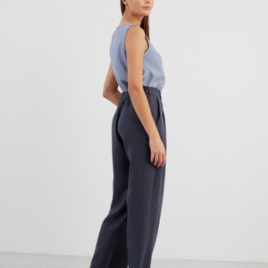 May include: A woman wearing a light blue sleeveless top and dark blue linen trousers. She is standing with her back to the camera and looking over her shoulder.