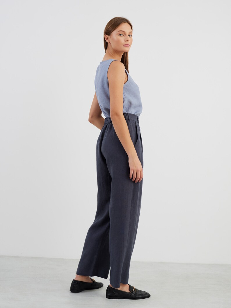 May include: A woman wearing a light blue sleeveless top and dark blue linen trousers. She is standing with her back to the camera and looking over her shoulder.
