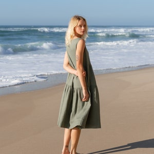 Puede incluir: Un vestido sin mangas de color verde oliva con falda escalonada. La persona lleva el vestido en una playa de arena, con el mar de fondo. La persona mira por encima del hombro hacia la c&aacute;mara.