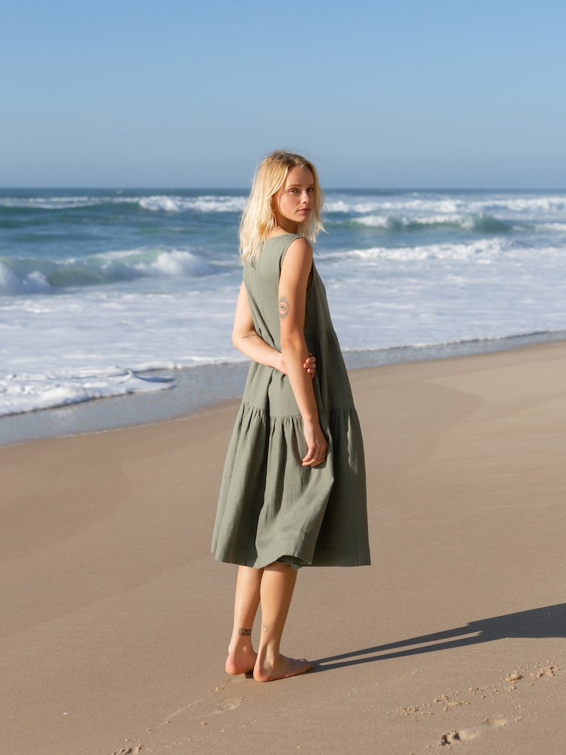 Puede incluir: Un vestido sin mangas de color verde oliva con falda escalonada. La persona lleva el vestido en una playa de arena, con el mar de fondo. La persona mira por encima del hombro hacia la c&aacute;mara.
