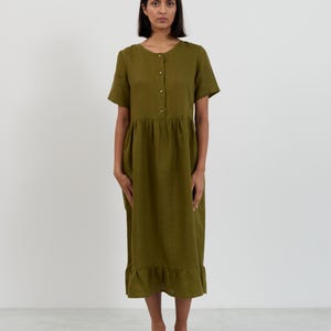 May include: Olive green midi dress with short sleeves, a button-down front, and a ruffled hem. The dress has a gathered waist and is made of a lightweight fabric. The model is wearing brown shoes.