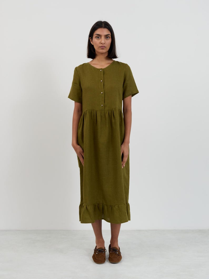 May include: Olive green midi dress with short sleeves, a button-down front, and a ruffled hem. The dress has a gathered waist and is made of a lightweight fabric. The model is wearing brown shoes.