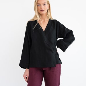 Op de afbeelding: Een zwart linnen wikkeltop met lange mouwen en een V-hals. Het top is in de taille gebonden met een ceintuur. Het model draagt ​​een bordeauxrode linnen broek.