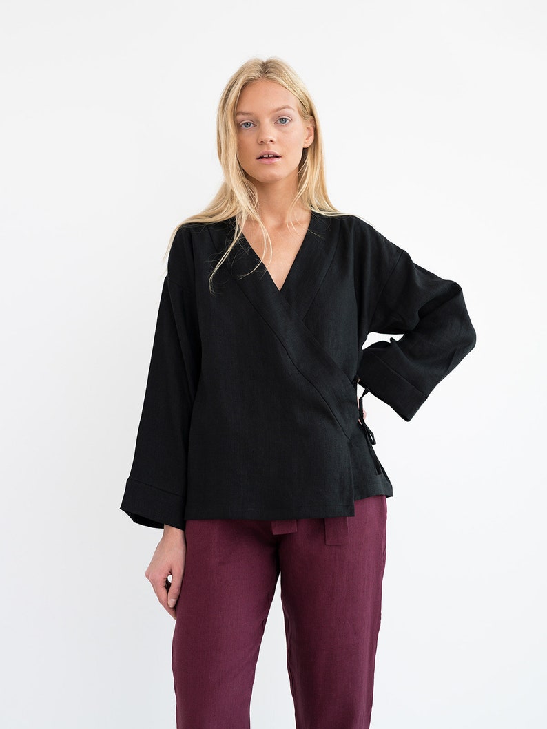 Op de afbeelding: Een zwart linnen wikkeltop met lange mouwen en een V-hals. Het top is in de taille gebonden met een ceintuur. Het model draagt ​​een bordeauxrode linnen broek.