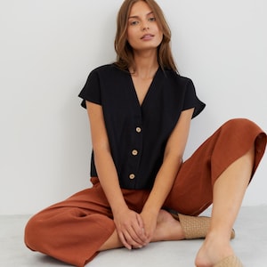 Puede incluir: Una mujer lleva una blusa negra de lino con botones, mangas cortas y cuello en V. También lleva pantalones anchos de color óxido y sandalias de cuero trenzado.