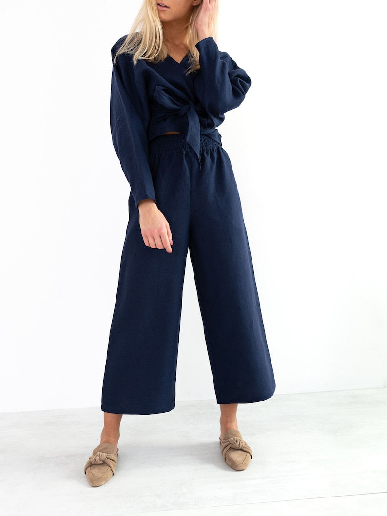 May include: A navy blue linen wrap top with a tie at the waist and matching wide-leg pants. The model is wearing the outfit with nude heeled mules.