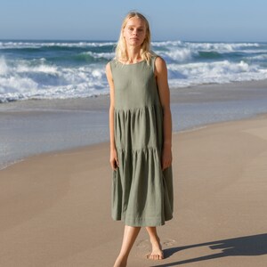 Puede incluir: Vestido midi sin mangas verde oliva con falda escalonada. Una persona est&aacute; de pie descalza en una playa de arena, con el oc&eacute;ano y las olas al fondo. El vestido tiene un dise&ntilde;o sencillo y relajado.
