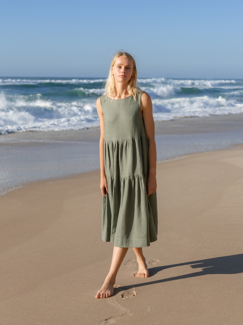 Puede incluir: Vestido midi sin mangas verde oliva con falda escalonada. Una persona est&aacute; de pie descalza en una playa de arena, con el oc&eacute;ano y las olas al fondo. El vestido tiene un dise&ntilde;o sencillo y relajado.
