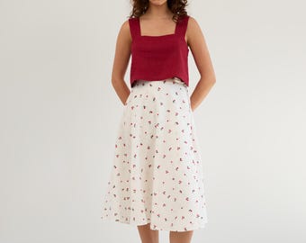 May include: A woman wearing a red sleeveless top and a white skirt with a small red floral pattern. She is wearing black loafers.