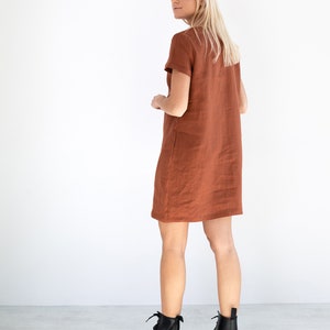 May include: A rust-colored linen dress with short sleeves and a straight silhouette. The dress is worn by a person with blonde hair and is paired with black ankle boots.