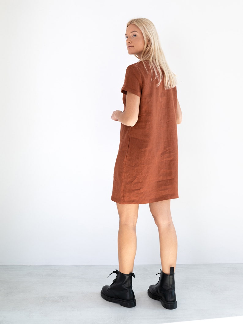May include: A rust-colored linen dress with short sleeves and a straight silhouette. The dress is worn by a person with blonde hair and is paired with black ankle boots.