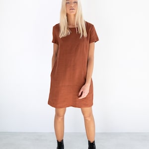 May include: A rust-colored linen dress with short sleeves and a square neckline. The dress is fitted through the bodice and falls to the mid-thigh. The model is wearing black ankle boots.