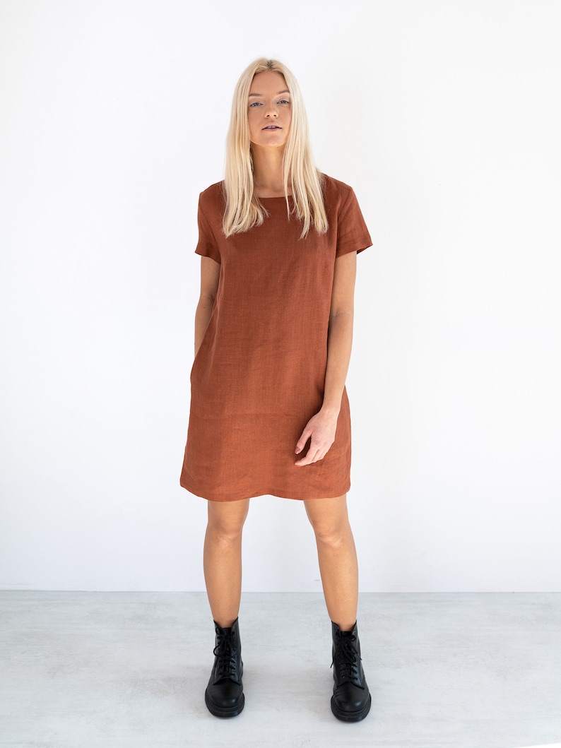 May include: A rust-colored linen dress with short sleeves and a square neckline. The dress is fitted through the bodice and falls to the mid-thigh. The model is wearing black ankle boots.