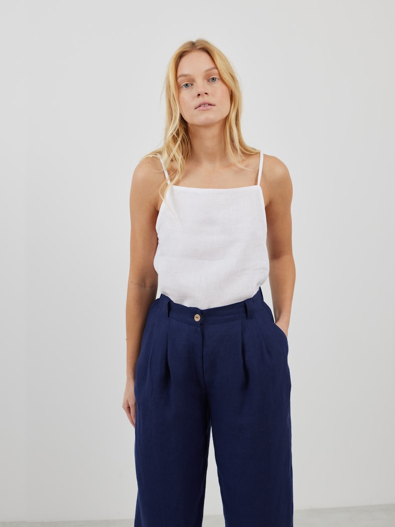 May include: A white linen tank top with thin straps and a square neckline, paired with dark blue wide-leg pants.