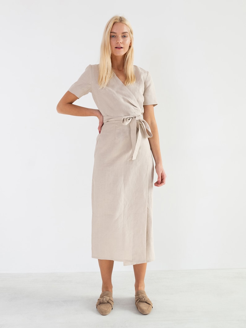 May include: A beige linen wrap dress with short sleeves and a tie belt. The dress is midi length and has a relaxed fit.