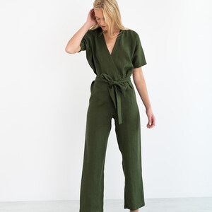 May include: A woman wearing a green linen jumpsuit with a wrap style bodice and a self-tie belt. The jumpsuit has short sleeves and wide legs.