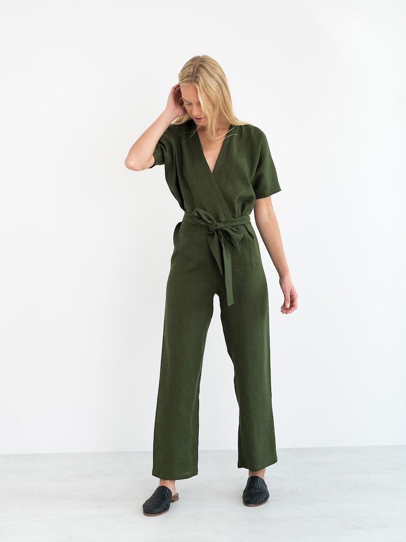 May include: A woman wearing a green linen jumpsuit with a wrap style bodice and a self-tie belt. The jumpsuit has short sleeves and wide legs.
