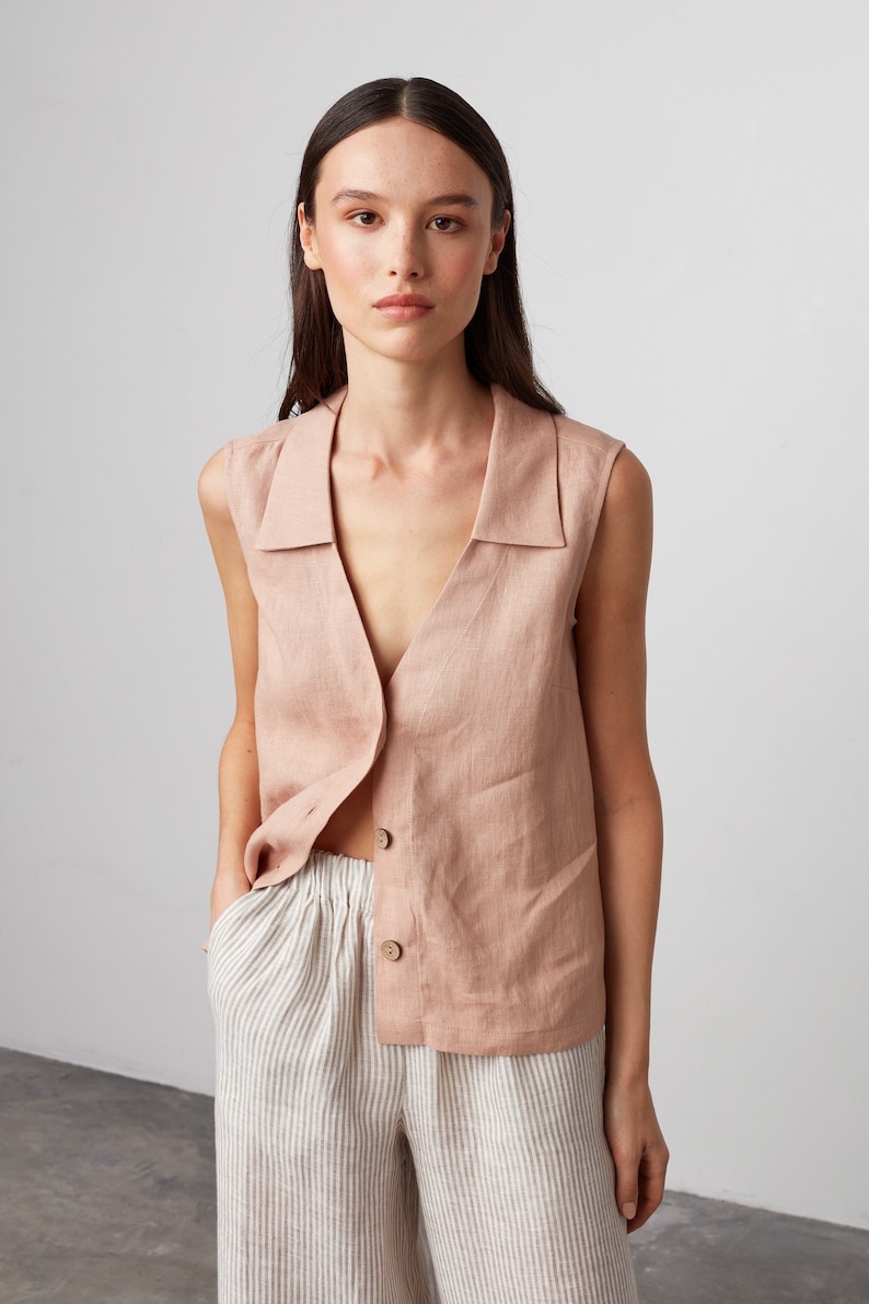 Pu&ograve; includere: Un top in lino senza maniche con un colletto e una chiusura con bottoni. Il top &egrave; di colore rosa chiaro e viene indossato sopra pantaloni in lino a righe.
