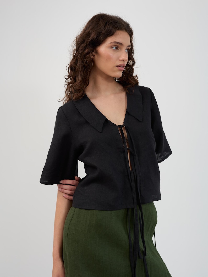 K&ouml;nnte beinhalten: Schwarzes kurz&auml;rmeliges Oberteil mit Kragen und Bindeband. Das Oberteil wird mit einer hoch taillierten, olivgr&uuml;nen Hose getragen. Das Outfit besteht aus einem leichten Stoff, und das Oberteil hat einen V-Ausschnitt. Das Model tr&auml;gt das Outfit in einem Studio.