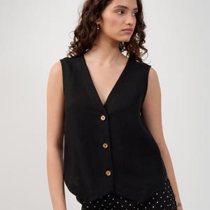 May include: A black sleeveless vest with a V-neck and wooden buttons. The vest is buttoned up and worn over a black and white polka dot skirt. The model is facing the camera with a neutral expression.