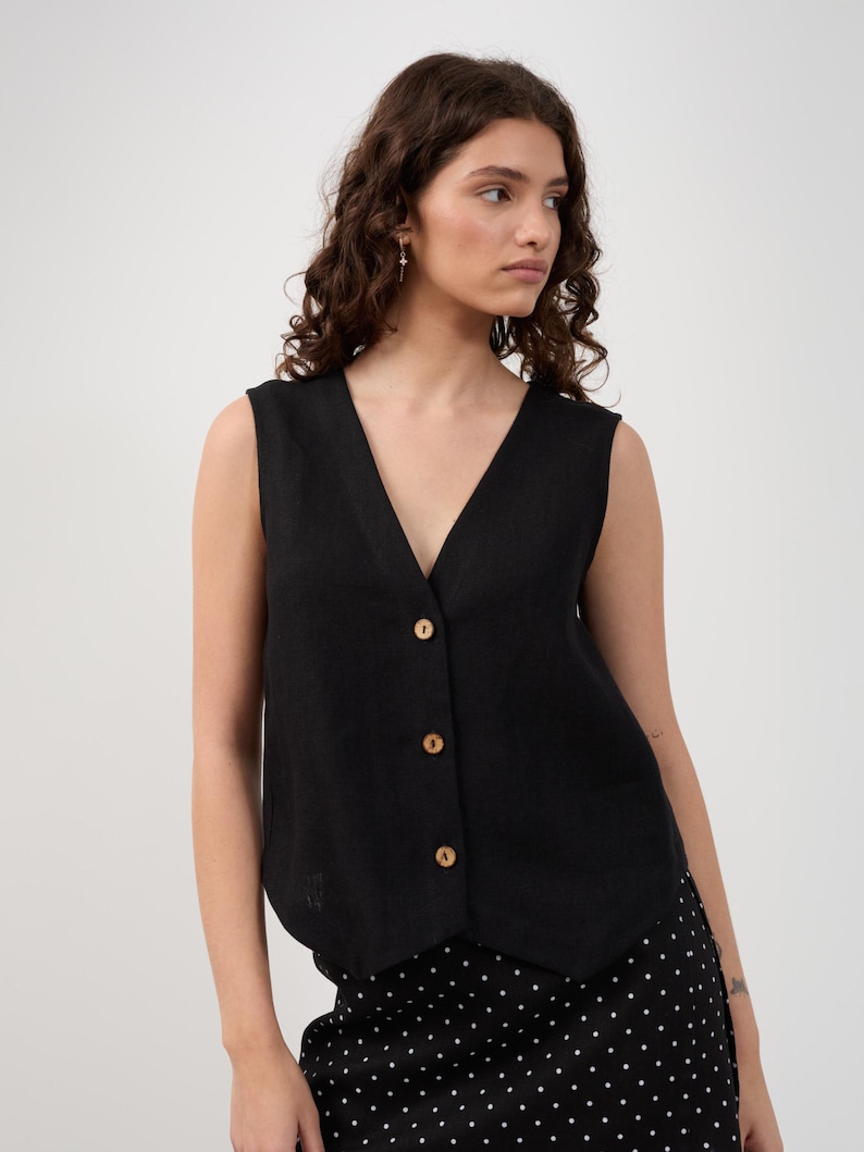 May include: A black sleeveless vest with a V-neck and wooden buttons. The vest is buttoned up and worn over a black and white polka dot skirt. The model is facing the camera with a neutral expression.