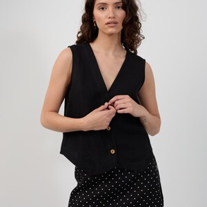 May include: A black sleeveless vest with a V-neck and wooden buttons. The vest is paired with a black skirt with white polka dots. The outfit is simple and stylish.