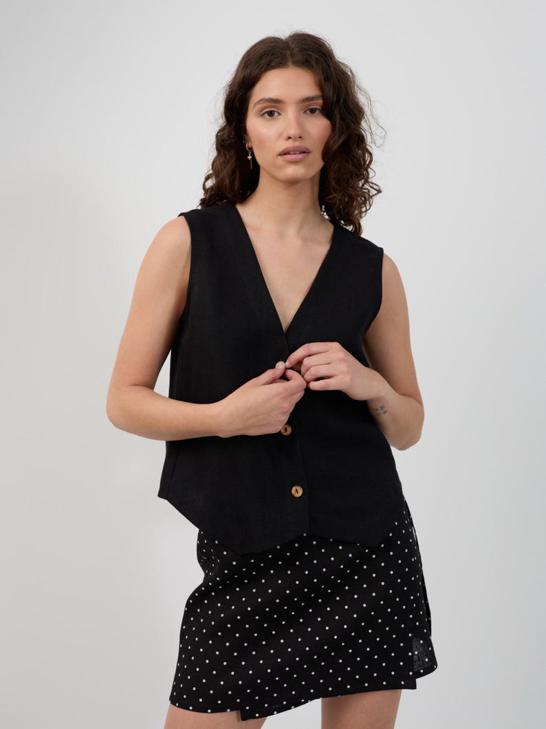 May include: A black sleeveless vest with a V-neck and wooden buttons. The vest is paired with a black skirt with white polka dots. The outfit is simple and stylish.