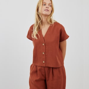 May include: A rust-colored linen top with a v-neck and button closure. The top has short sleeves and is paired with matching rust-colored linen pants.